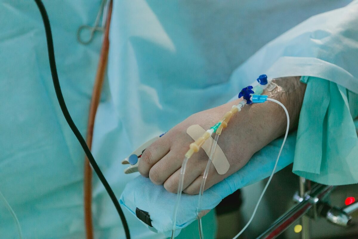 A patient’s hand with intravenous lines and monitoring clips rests on a padded surface, surrounded by medical equipment and blue surgical drapes.