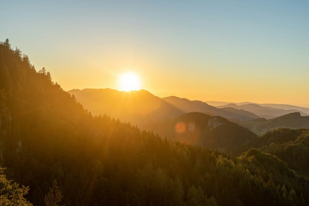 The sun sets over a range of forested mountains, casting a golden light across the landscape under a clear sky.