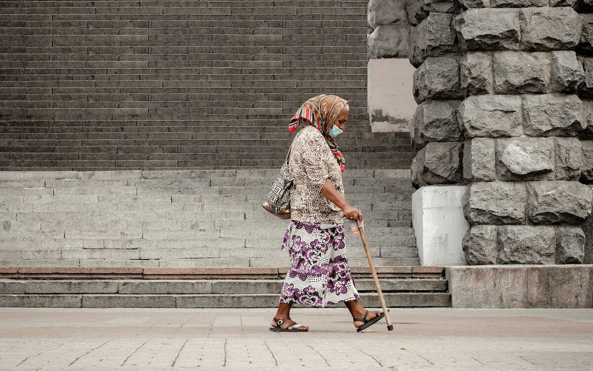 An older woman wearing a headscarf and face mask walks with a cane past a large stone wall and steps.