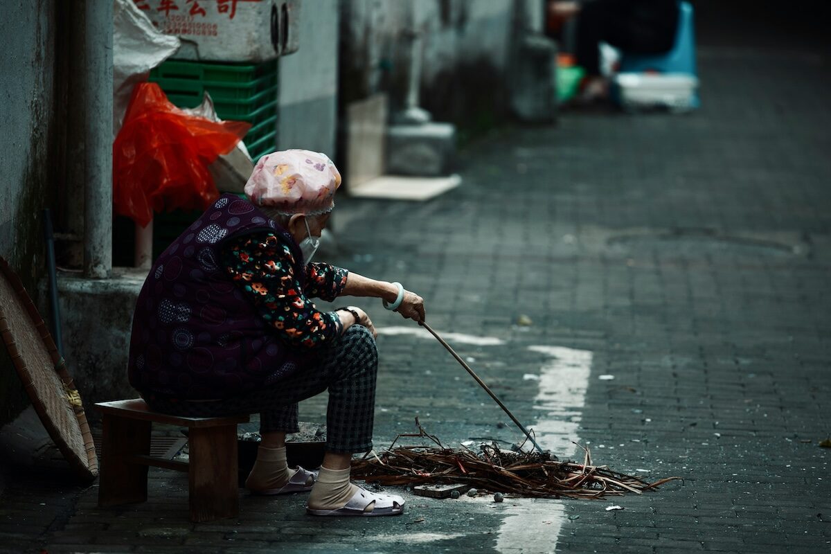 An elderly person sits on a small stool beside an alley, sorting through a pile of sticks with a stick in hand.