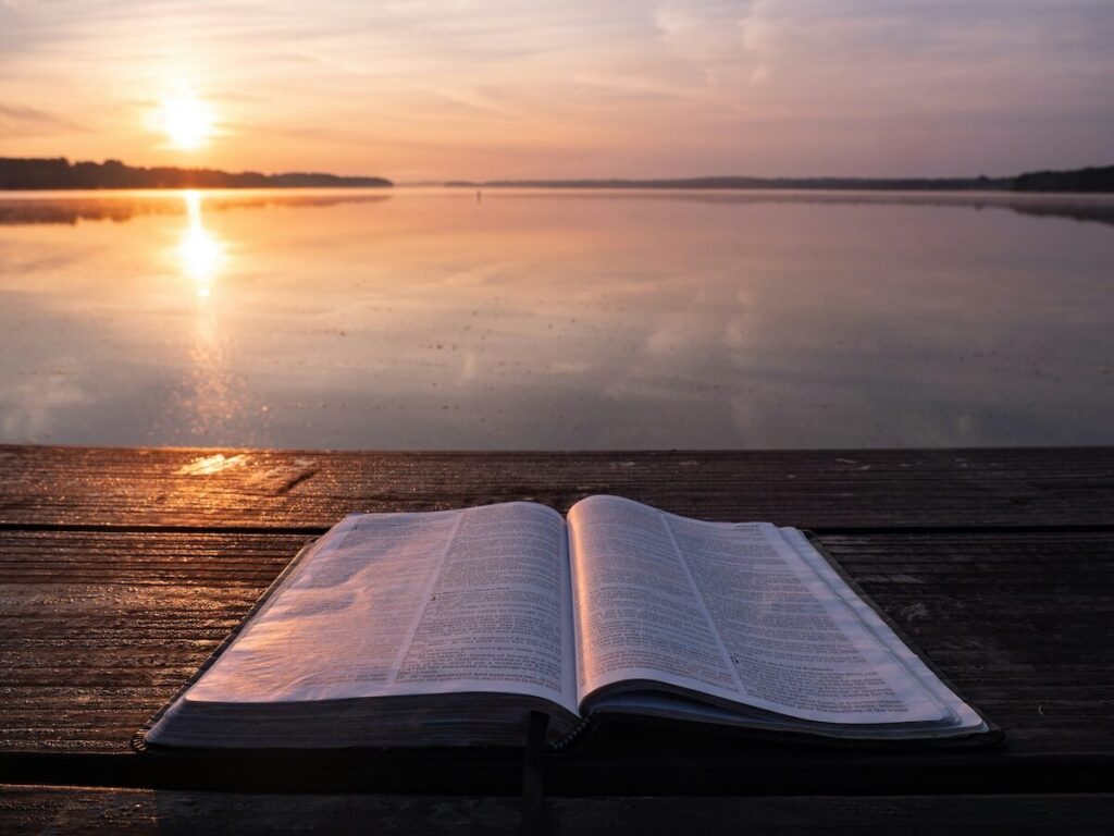 An open book rests on a wooden surface by a calm lake at sunset, with the sun low on the horizon and its reflection visible in the water.
