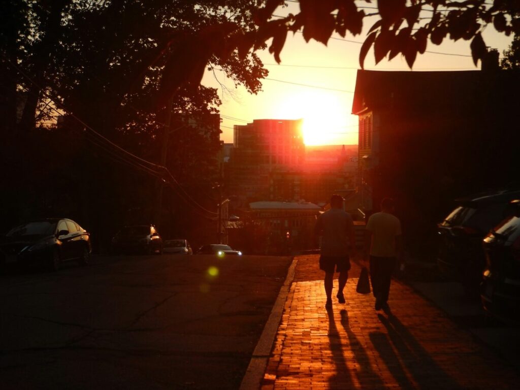 Two people walk on a sidewalk at sunset, with warm sunlight illuminating the street and casting long shadows.