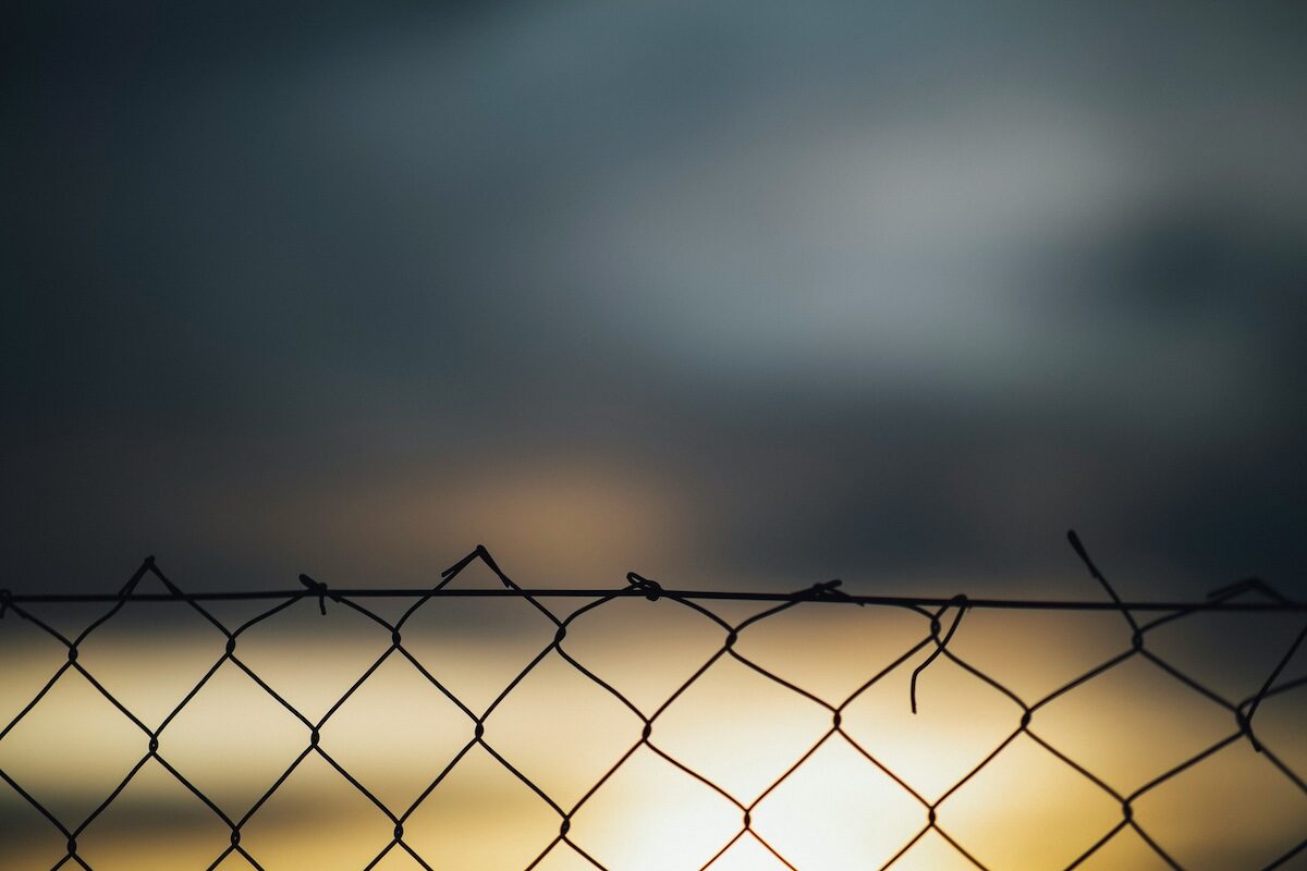 A close-up view of a wire fence with a blurred, cloudy sky and soft light in the background.