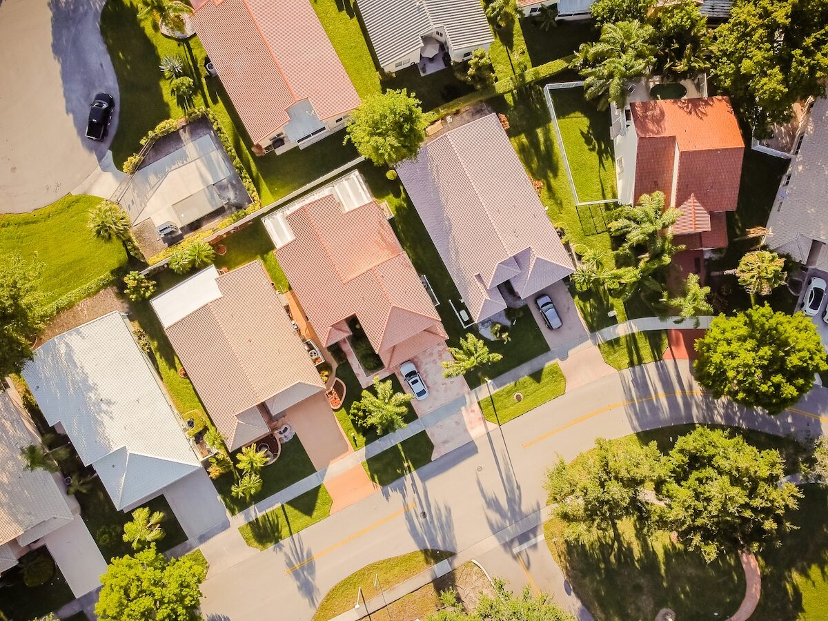 Aerial view of a suburban neighborhood showing several houses with driveways, lawns, and trees lining the street under sunlight.