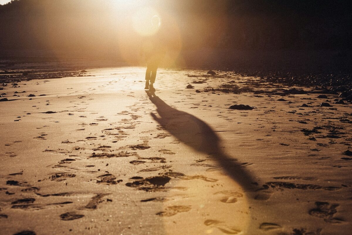 A person walks along a sandy beach at sunset, casting a long shadow across the sand with footprints visible.