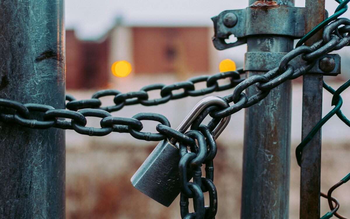 Close-up of a metal gate secured with a chain and padlock, with blurred buildings and lights in the background.