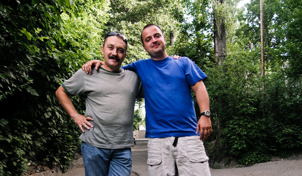 Two men pose for a portrait in front of some tall trees.