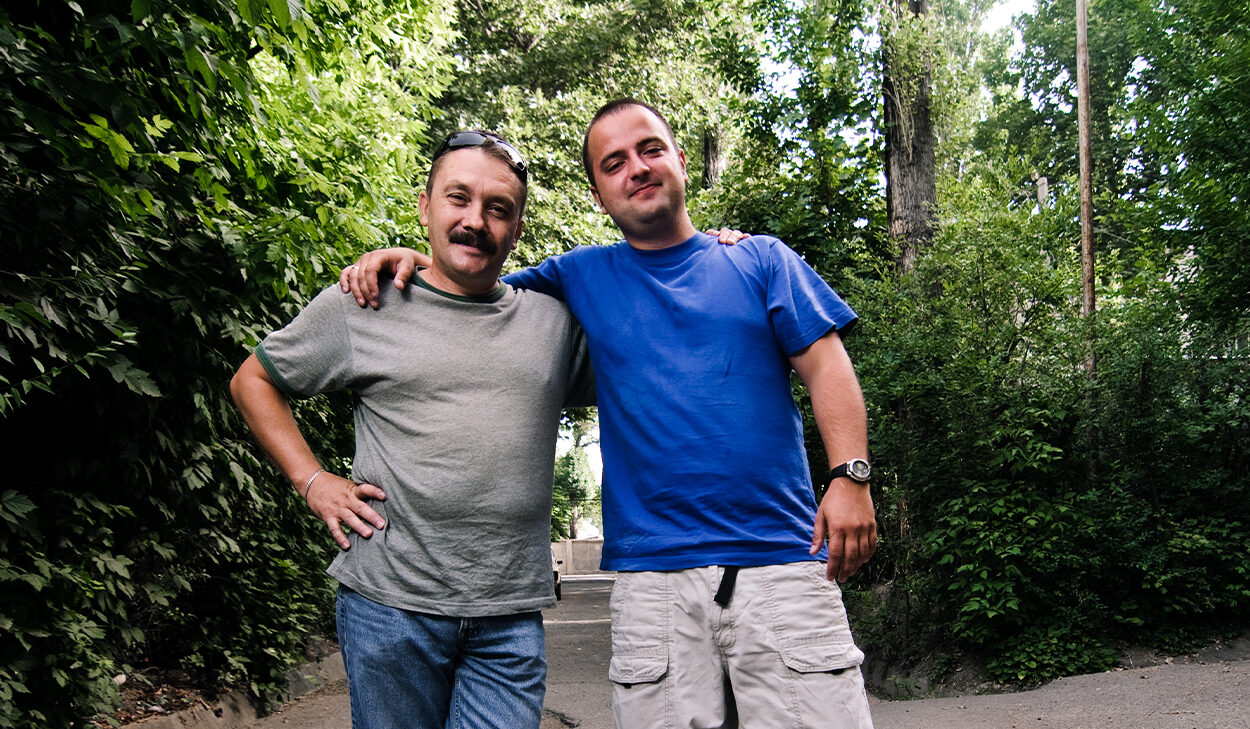 Two men pose for a portrait in front of some tall trees.