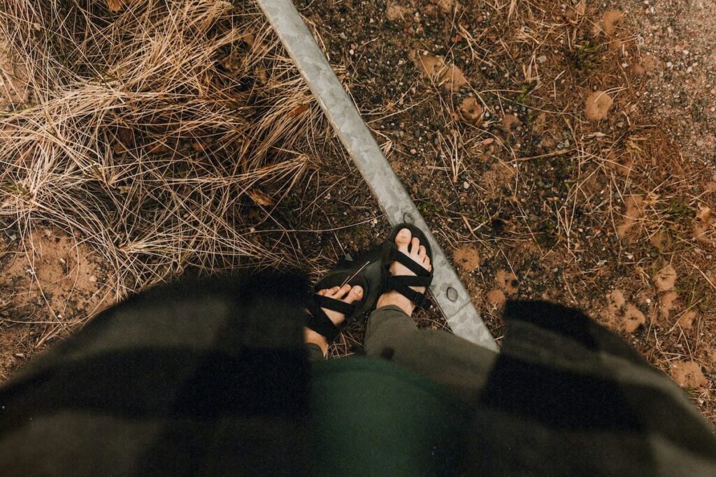 Person wearing sandals and a plaid shirt stands on or near a metal rail, with dry grass and dirt visible on the ground.
