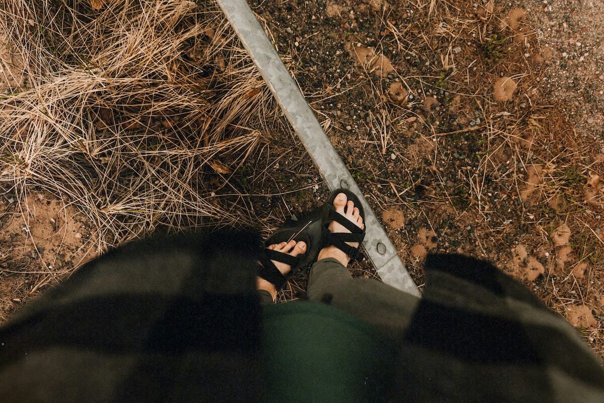 Person wearing sandals and a plaid shirt stands on or near a metal rail, with dry grass and dirt visible on the ground.