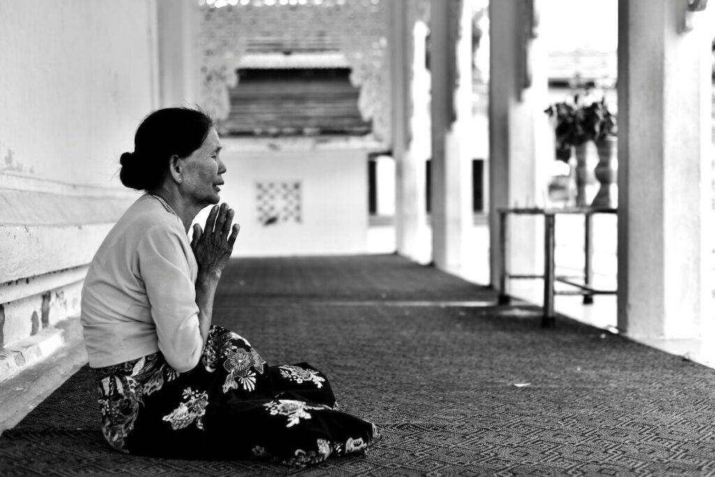 A woman sits on the floor indoors with her hands pressed together in prayer, facing forward, in a quiet, possibly religious setting.