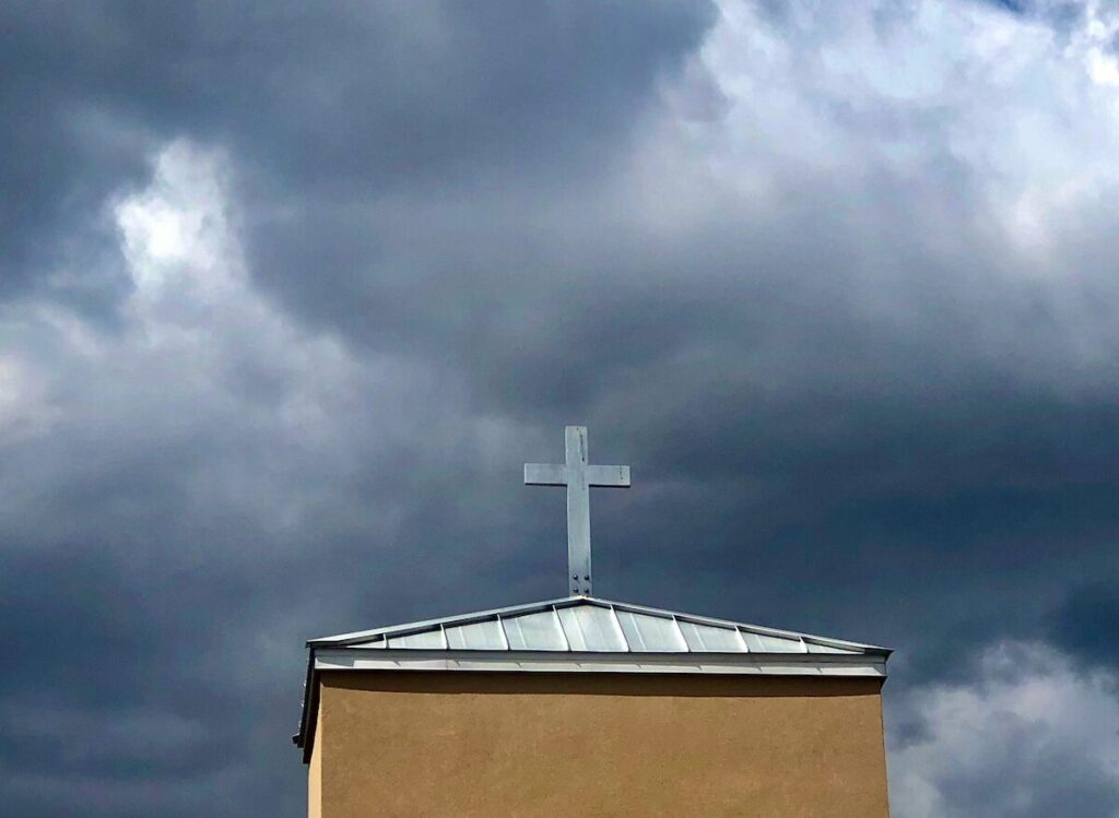 A metal cross stands on the roof of a beige building under a cloudy, dark sky.