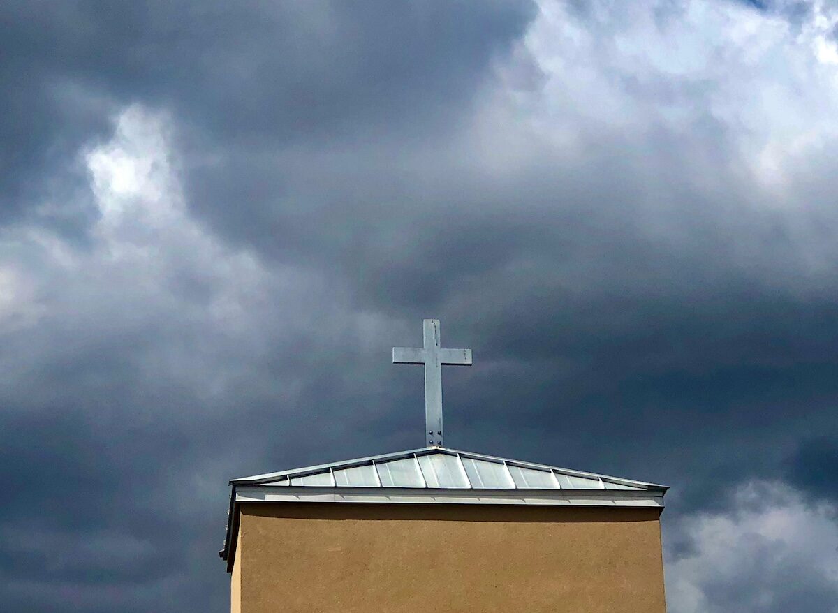 A metal cross stands on the roof of a beige building under a cloudy, dark sky.