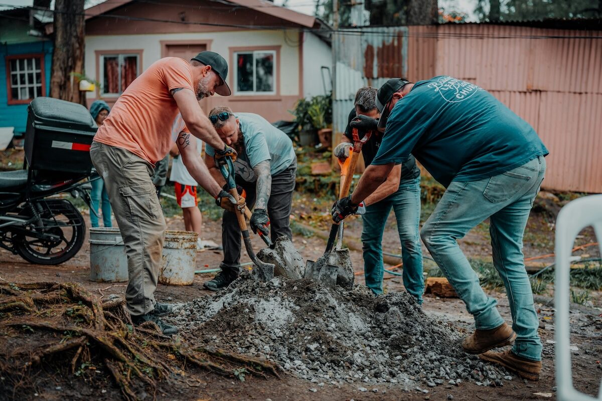 Four people work together shoveling gravel or cement mixture outdoors, with houses and a motorcycle visible in the background.