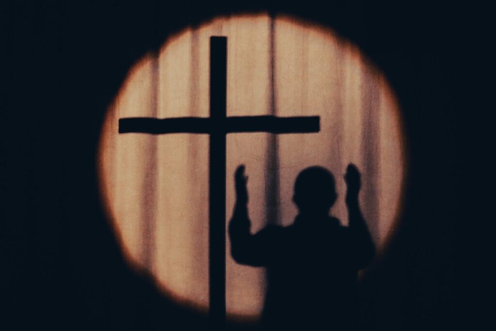 A silhouette of a person with raised hands stands next to a large cross, both cast in shadow against a circular spotlight on a curtain.