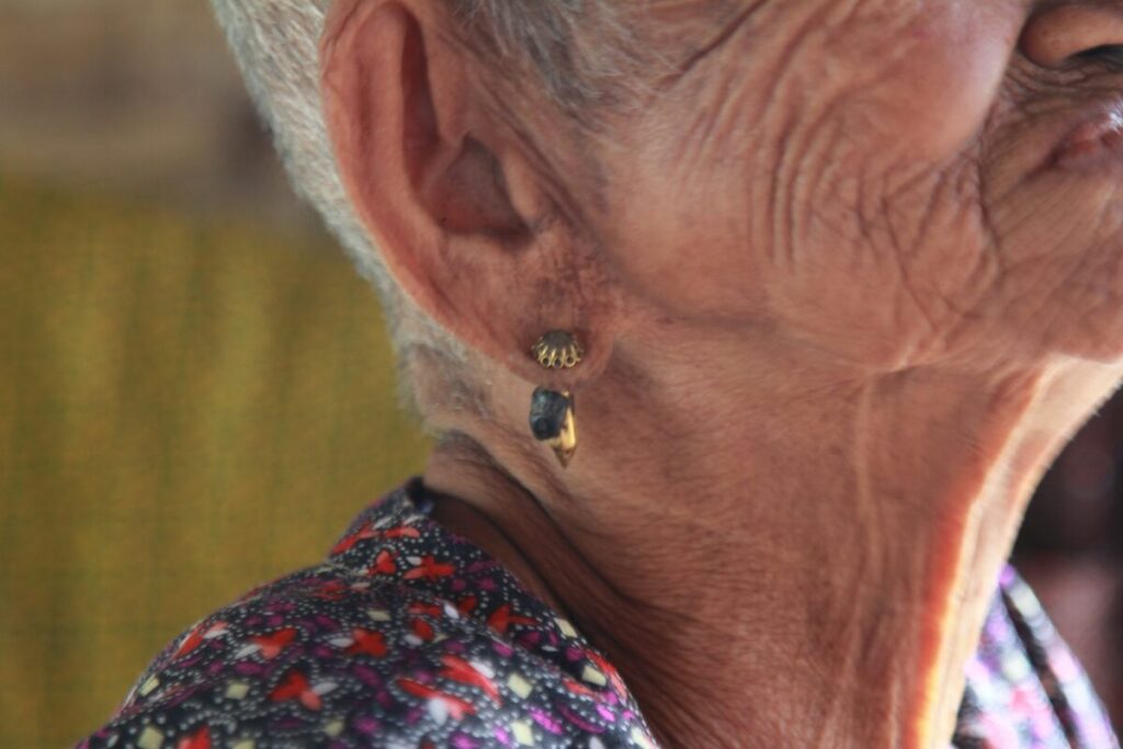 Close-up of an elderly persons neck, jaw, and ear, showing wrinkles, a short gray haircut, and a dangling earring. The person is wearing a patterned top.