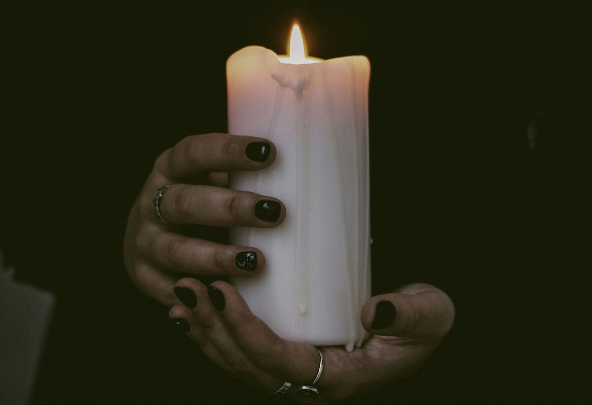 A person with black nail polish and rings holds a lit white candle against a dark background.