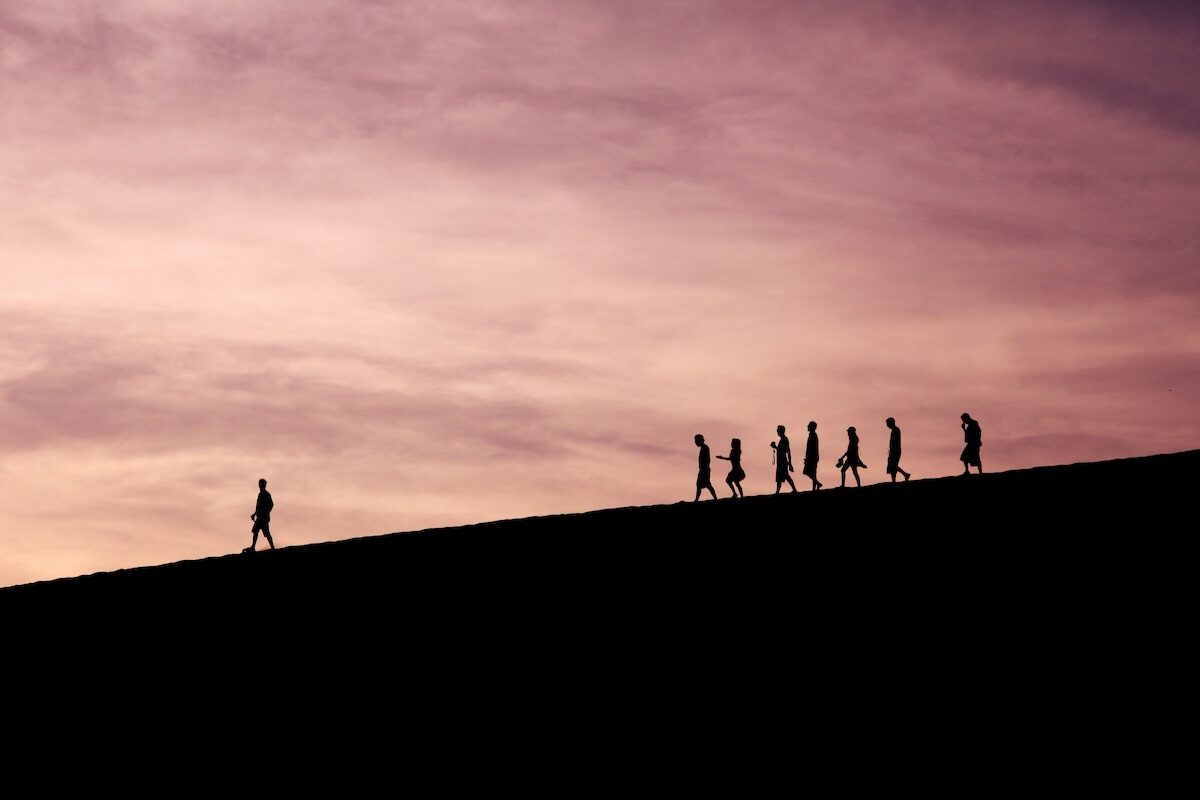 Seven people are silhouetted, walking in a line along a sloped hill at sunset under a pink and purple sky.