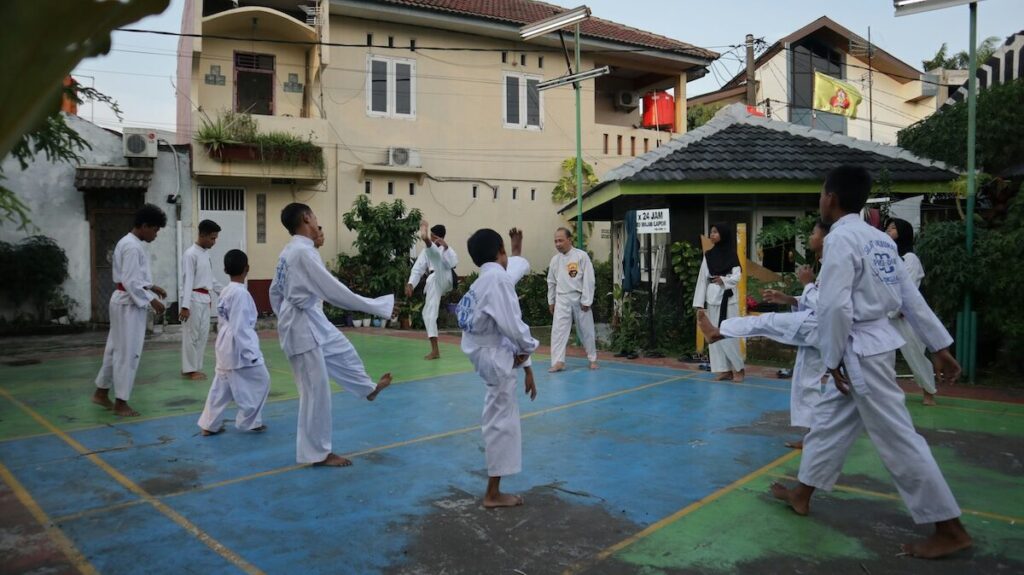 A group of people in white martial arts uniforms practice together on an outdoor court in a residential area during daylight.