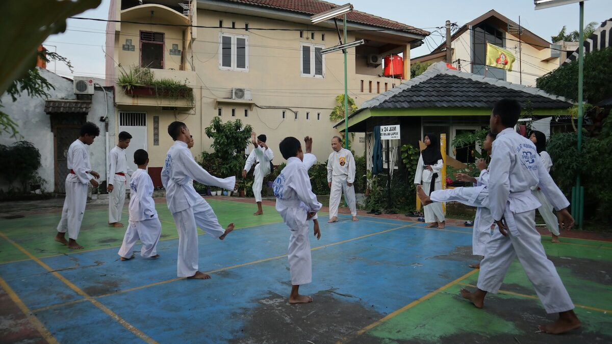 A group of people in white martial arts uniforms practice together on an outdoor court in a residential area during daylight.