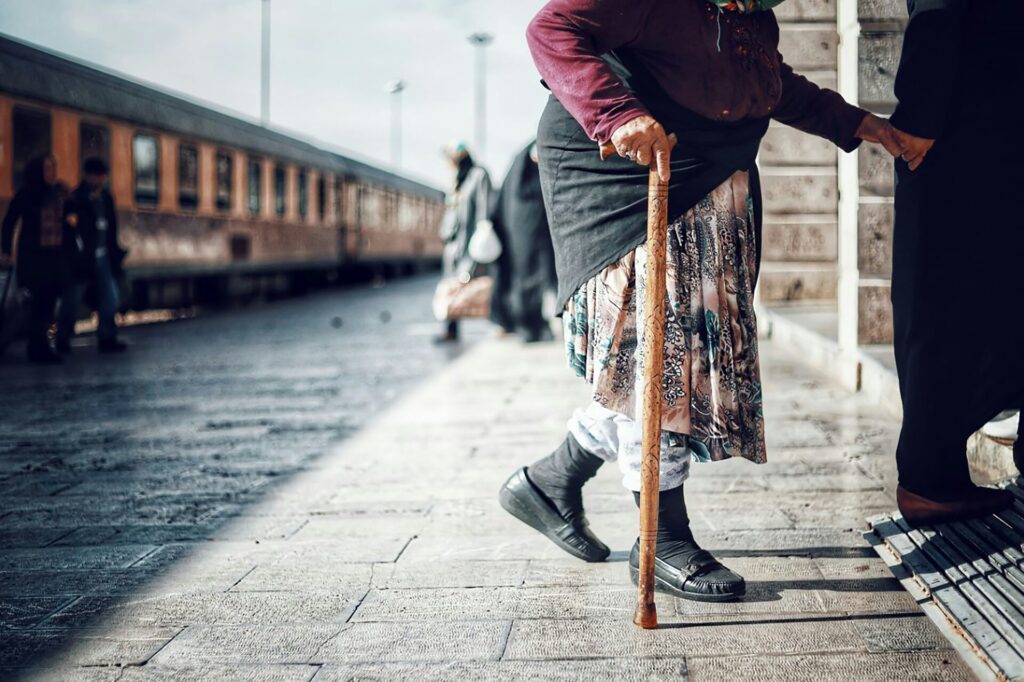 An older person with a cane stands on a train platform next to a brick wall, wearing a skirt, sweater, and headscarf. A train and several people are visible in the background.