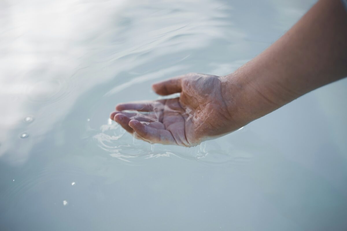 A hand is partially submerged in clear water, with fingers slightly curved and droplets visible on the skin.