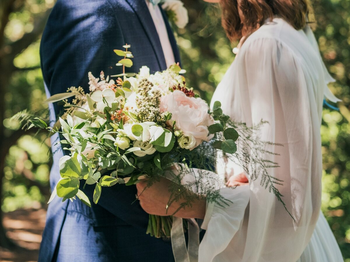 A close-up of a couple at an outdoor wedding, with the bride holding a bouquet of white and pink flowers and greenery.
