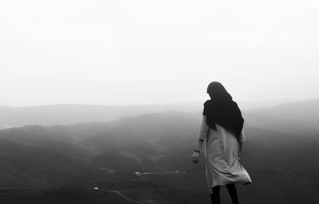 A person in a long coat stands on a hill, looking out over a misty, mountainous landscape.
