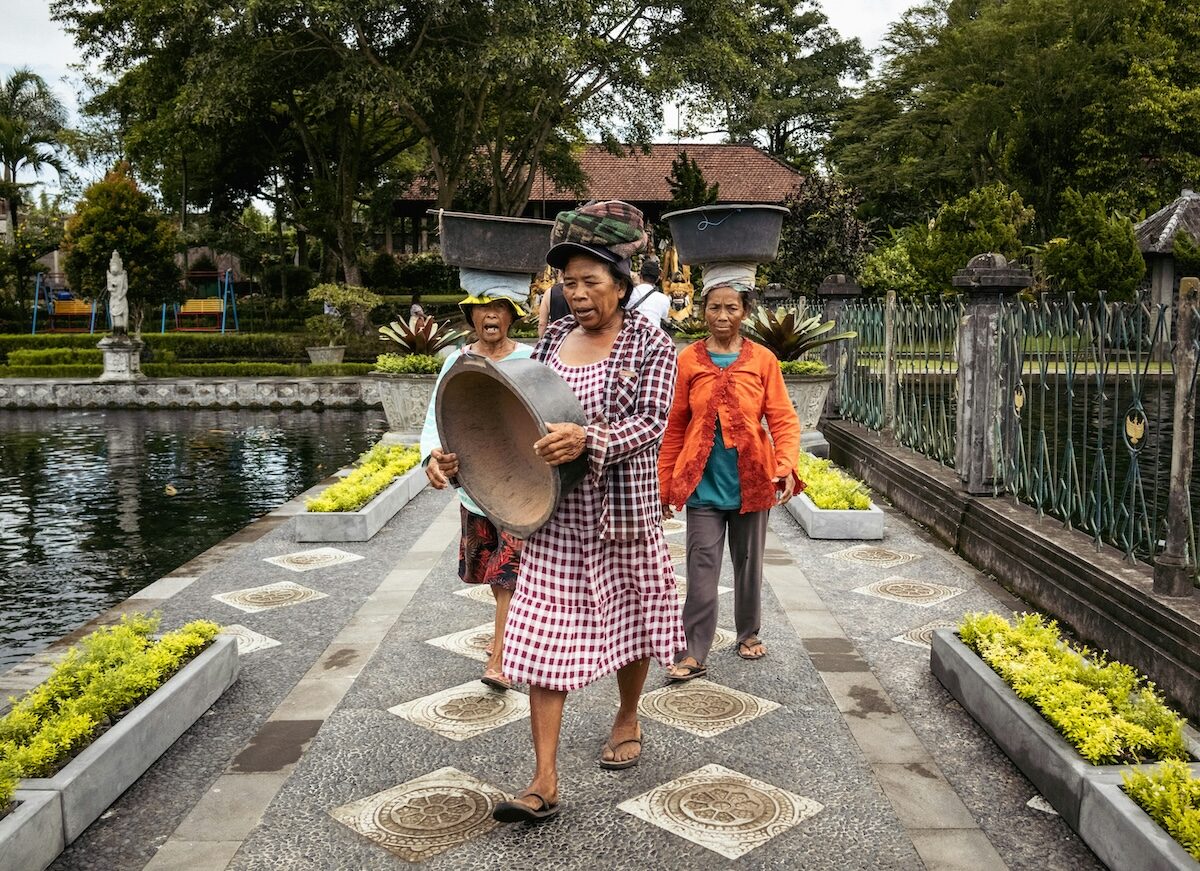Four women walk along a garden path by a pond, with one woman in front carrying a large round tray. Trees and a building are visible in the background.