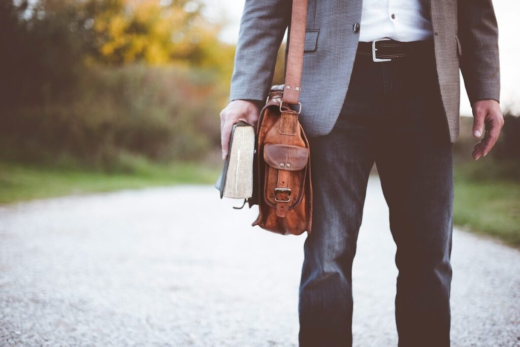 A person in a suit stands on a gravel path holding a book and a brown leather shoulder bag.