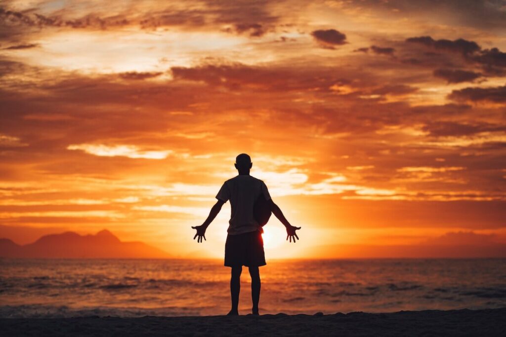 A person stands on a beach at sunset with arms outstretched, silhouetted against an orange and yellow sky over the ocean.