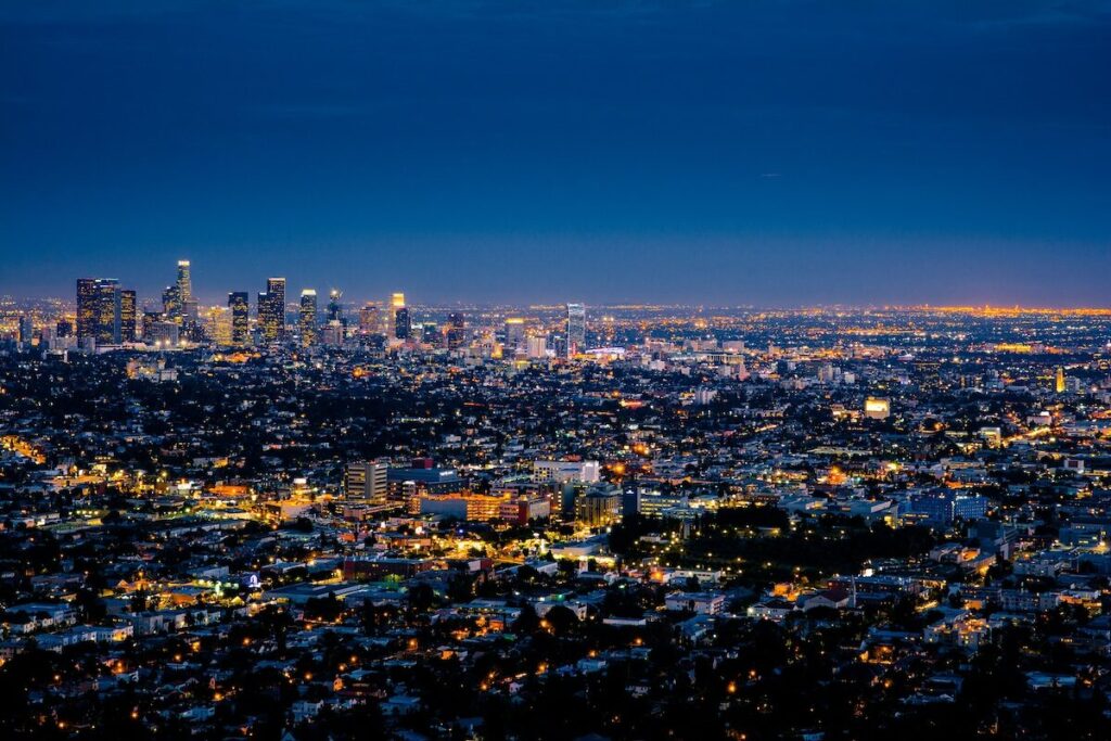 Aerial view of a cityscape at dusk, with illuminated buildings and streets, and a brightly lit downtown skyline in the distance.