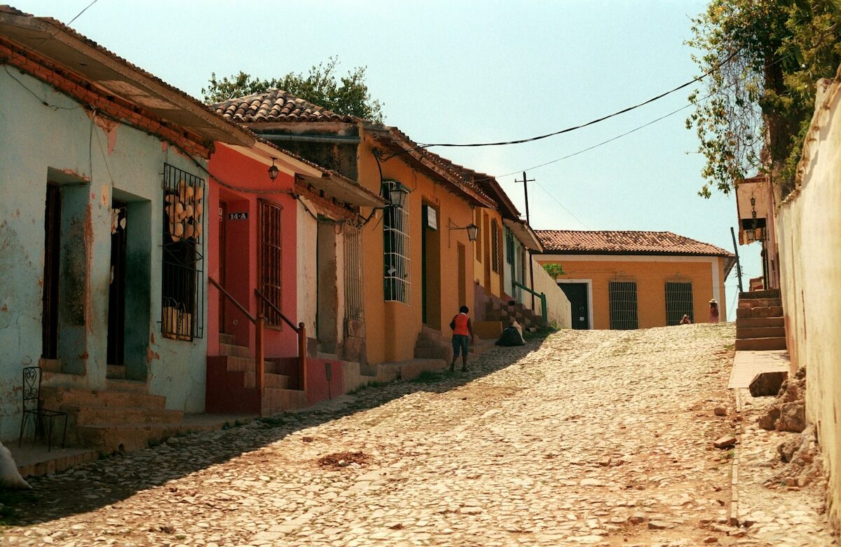 A narrow, cobblestone street lined with colorful, weathered buildings on one side and a stone wall on the other, with a person walking uphill in the distance.