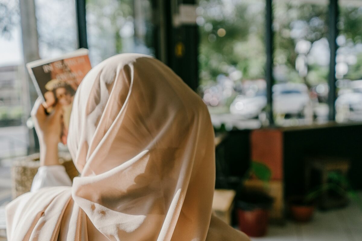 A person wearing a light-colored headscarf is seen from behind while holding up a magazine inside a room with large windows.