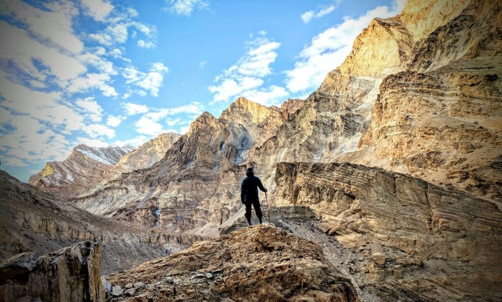 A person stands on a rocky outcrop amid rugged, sunlit mountains under a partly cloudy sky.
