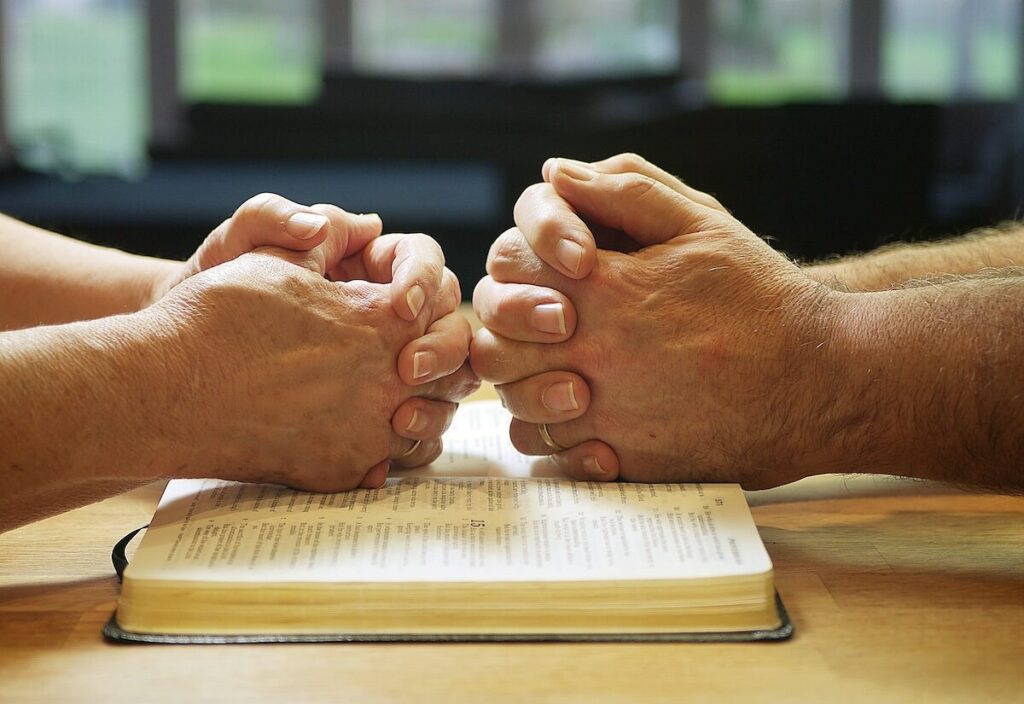 Two pairs of hands are clasped in prayer over an open book, likely a Bible, on a wooden table with a blurred background.
