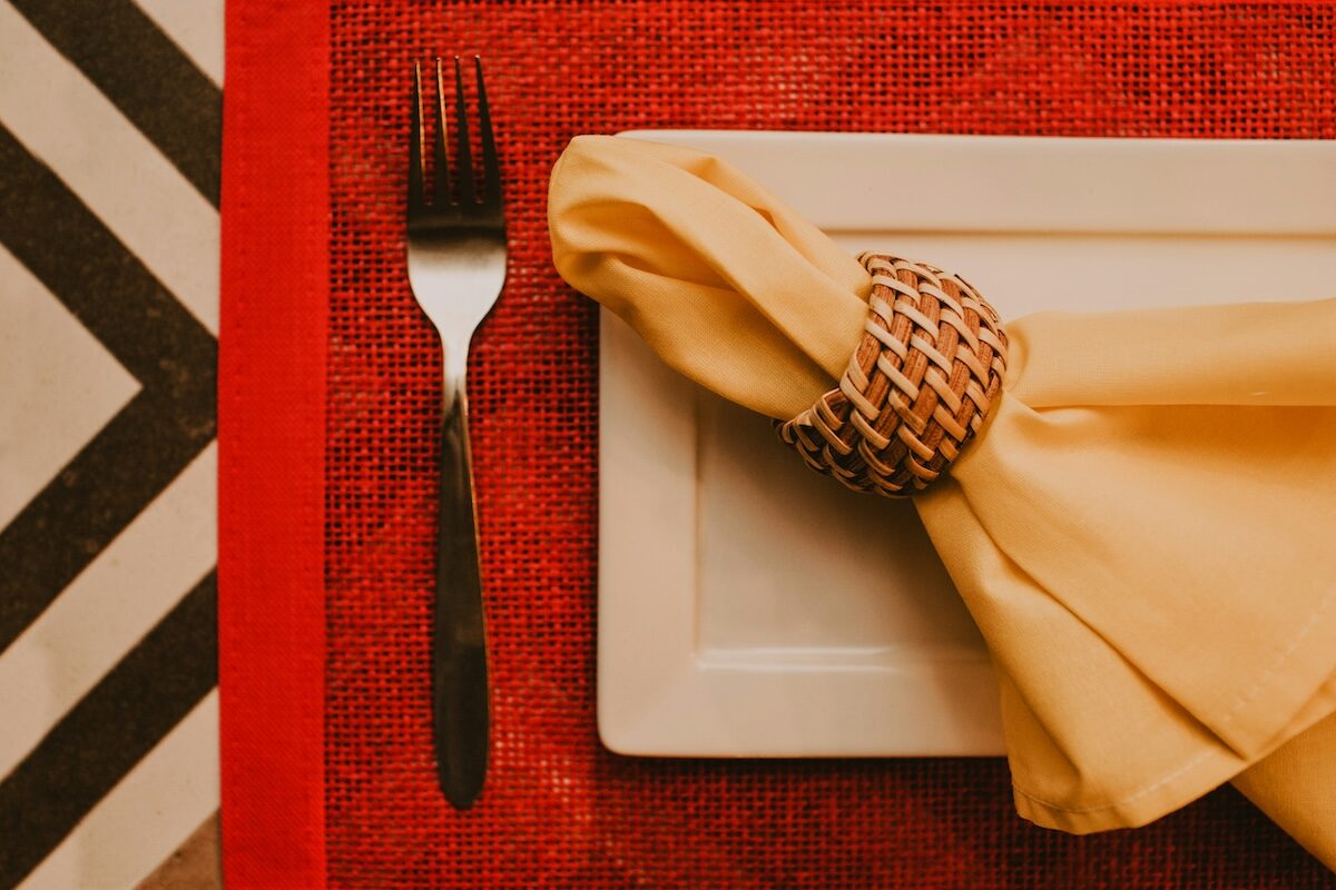 A fork and a rectangular white plate with a folded yellow napkin in a decorative ring are set on a red placemat with a black and white zigzag pattern beside it.