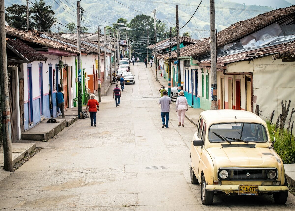 A narrow street in a small town with colorful houses, a yellow vintage car parked on the right, and several people walking along the sidewalk.