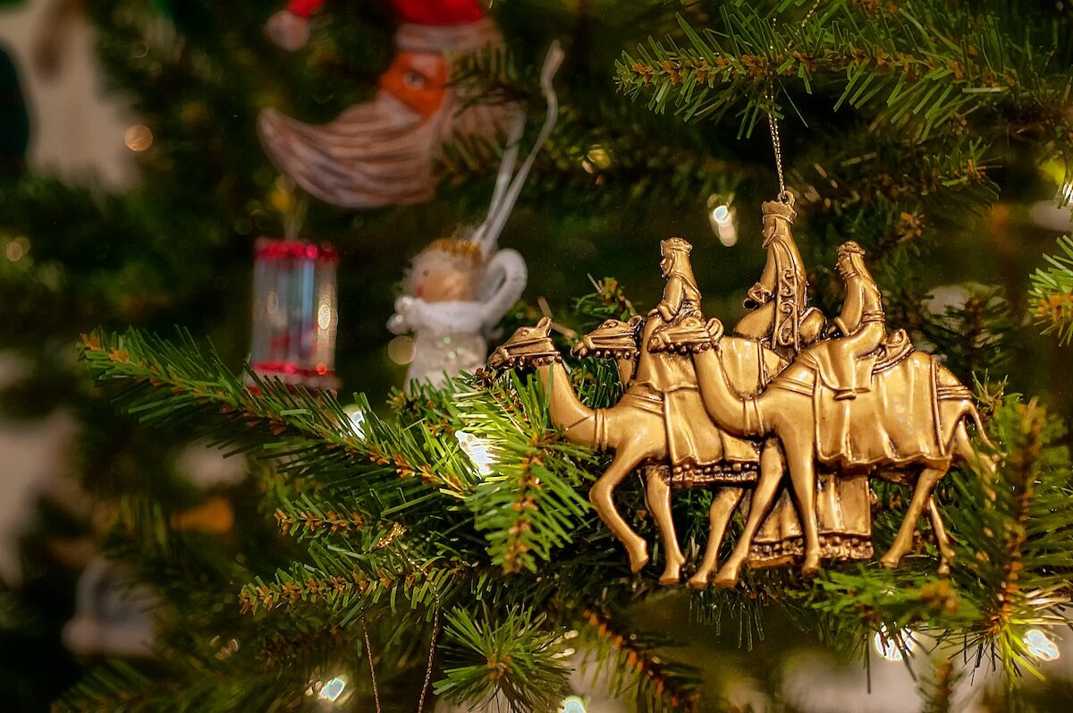 A gold ornament of the Three Wise Men riding camels hangs on a Christmas tree, surrounded by green branches and other decorations.