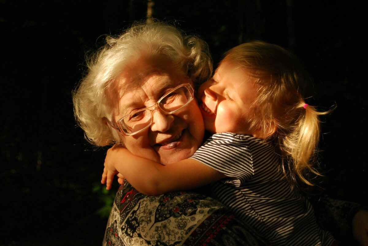 An elderly woman with glasses smiles as a young child hugs and kisses her cheek in a warmly lit setting.