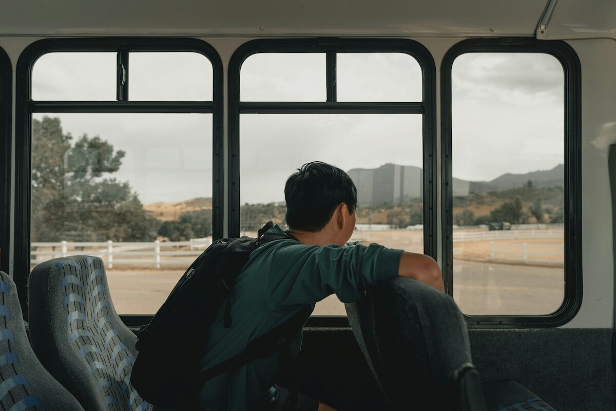 A person with a backpack sits alone on a bus, looking out the window at a rural landscape with mountains in the distance.