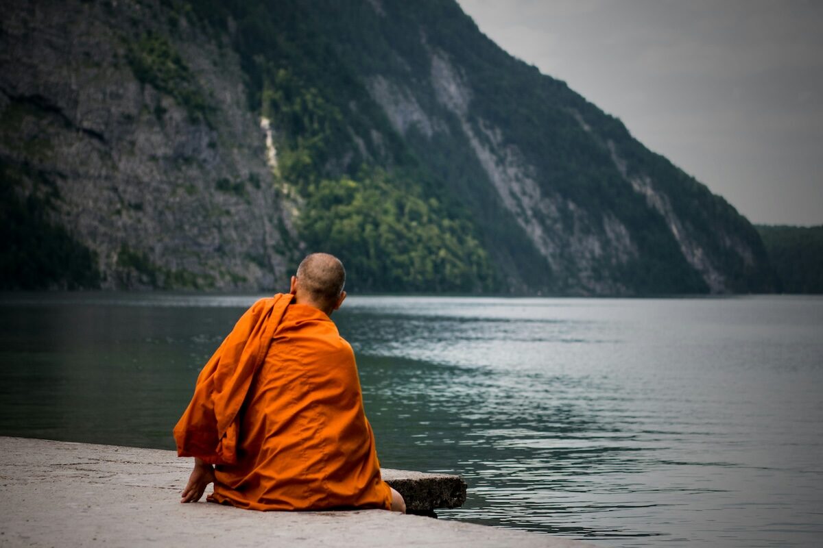 A person wearing an orange robe sits on the edge of a body of water, facing away toward a large, forested mountain.
