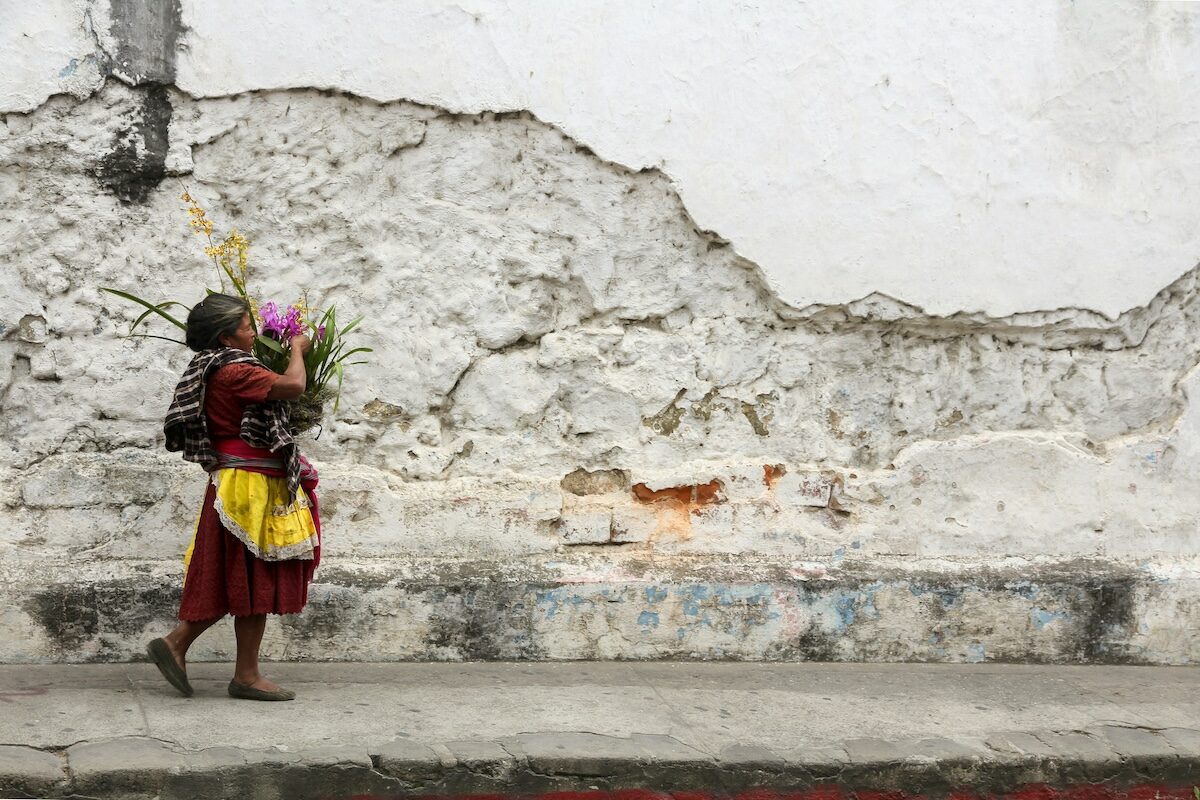 A woman in traditional clothing walks along a cracked, weathered wall carrying a child and a bundle of flowers.