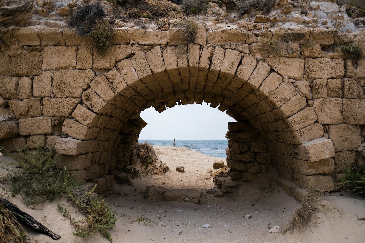 Stone archway with missing blocks frames a view of sand, sea, and sky, with a person standing in the distance near the water.