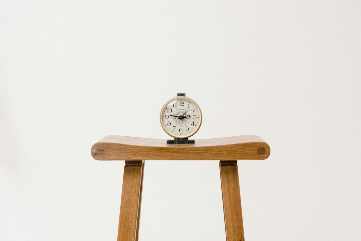 A small analog alarm clock sits on top of a wooden stool against a plain white background.