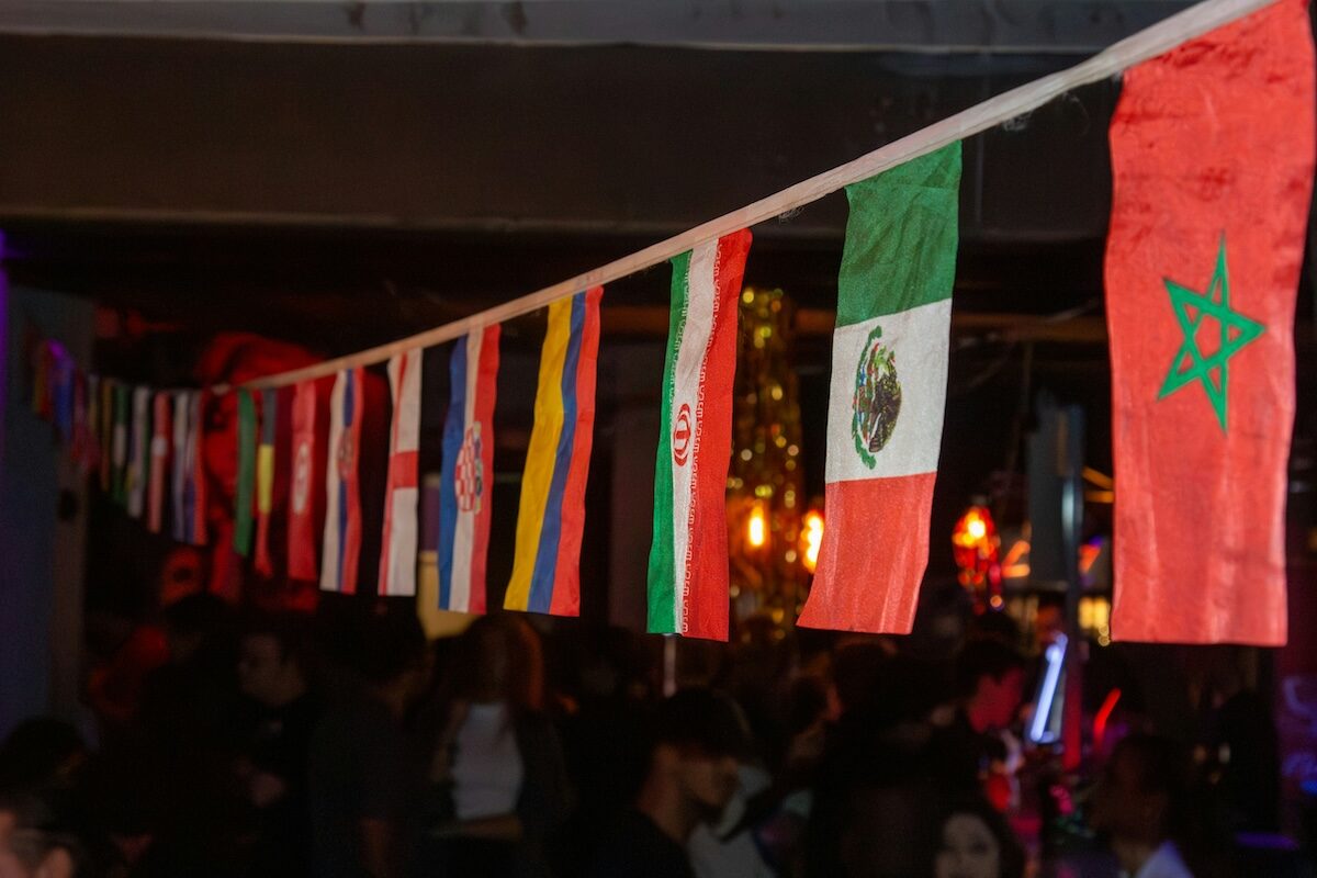 A string of various international flags hangs indoors above a crowd of people in a dimly lit space.