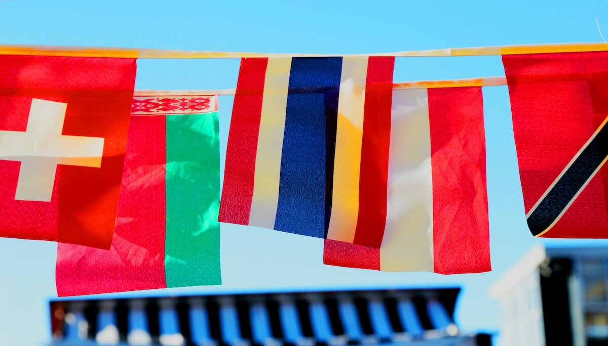 A row of international flags, including those of Switzerland, Belarus, Thailand, Austria, and Trinidad and Tobago, hanging against a clear blue sky.