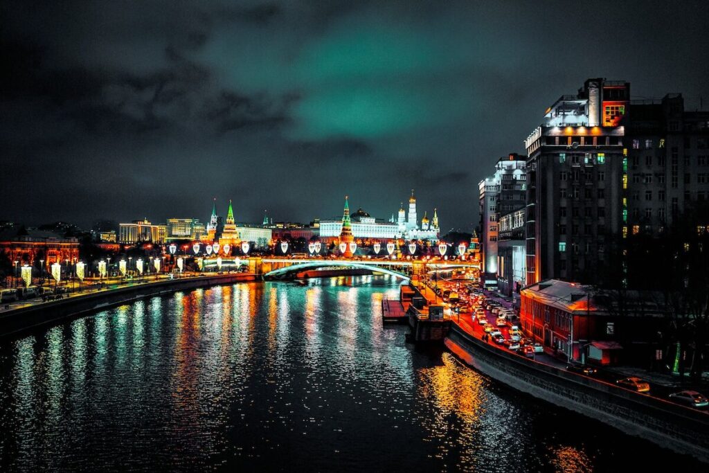 Night view of a cityscape with illuminated buildings and bridge reflected in a river, featuring traffic and cloudy skies.