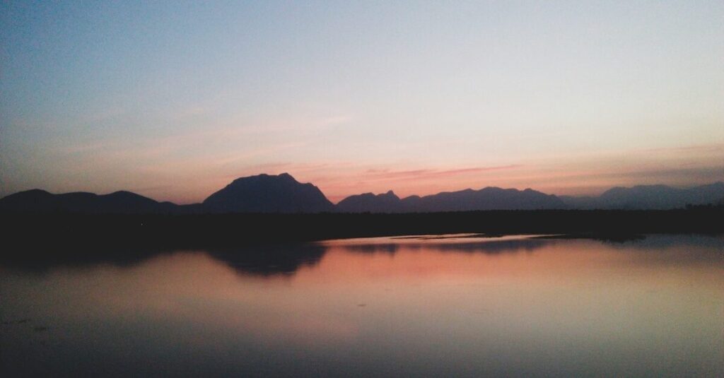 A calm lake reflects distant mountains and a pastel-colored sky at sunset.