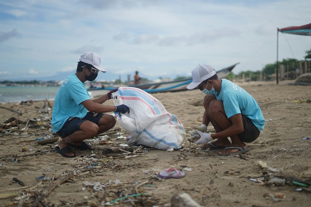 Two people wearing masks and gloves collect trash and debris into a large bag on a sandy beach, with the sea and boats visible in the background.